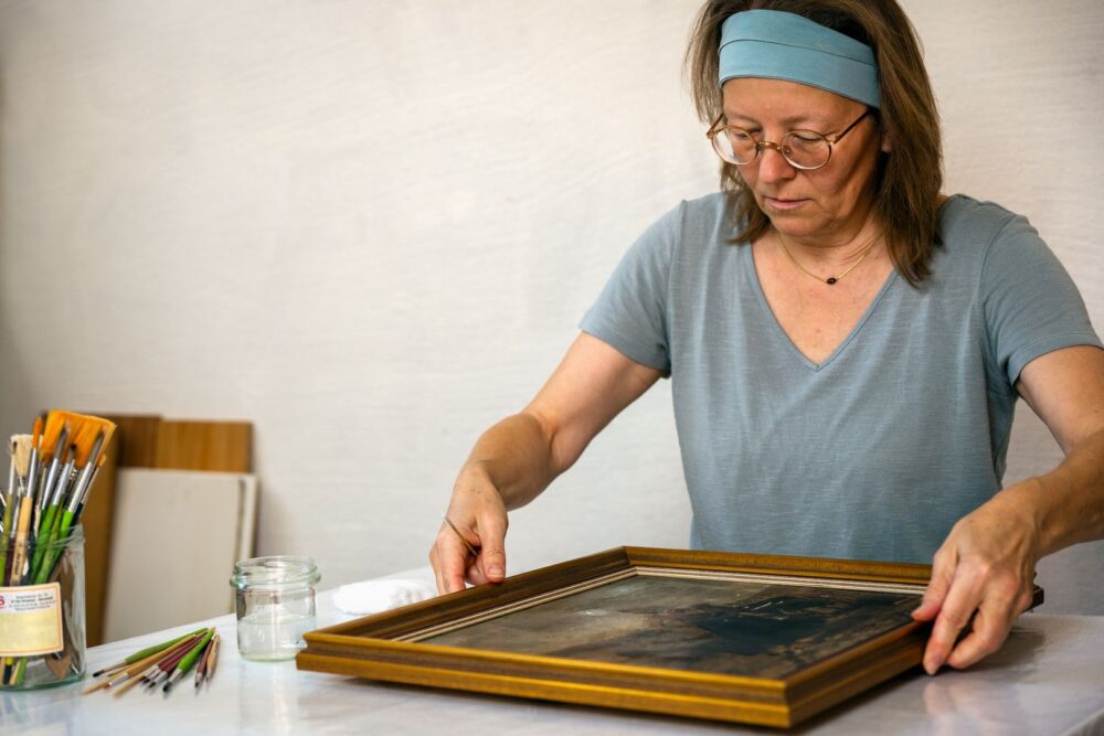 Restauratorin untersucht gerahmtes Gemälde mit dunkler Malschicht am Arbeitstisch, umgeben von Pinseln,  Foto © Stefan Trost