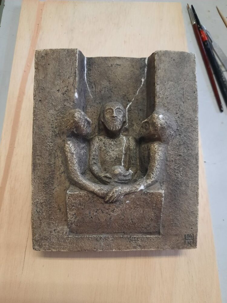 Das kleine Relief aus Kunststein war zerbrochen und wurde wieder zusammengefügt. Die weiß gekitteten Risse wurden anschließend im Umgebungston retuschiert.